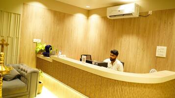 A reception desk with a person seated behind it, a wall-mounted air conditioner, and a sofa with a decorative lamp.