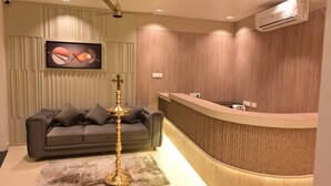 A reception area with a curved desk, a grey sofa, and a decorative lamp. - Saba's Courtyard (Colachel)