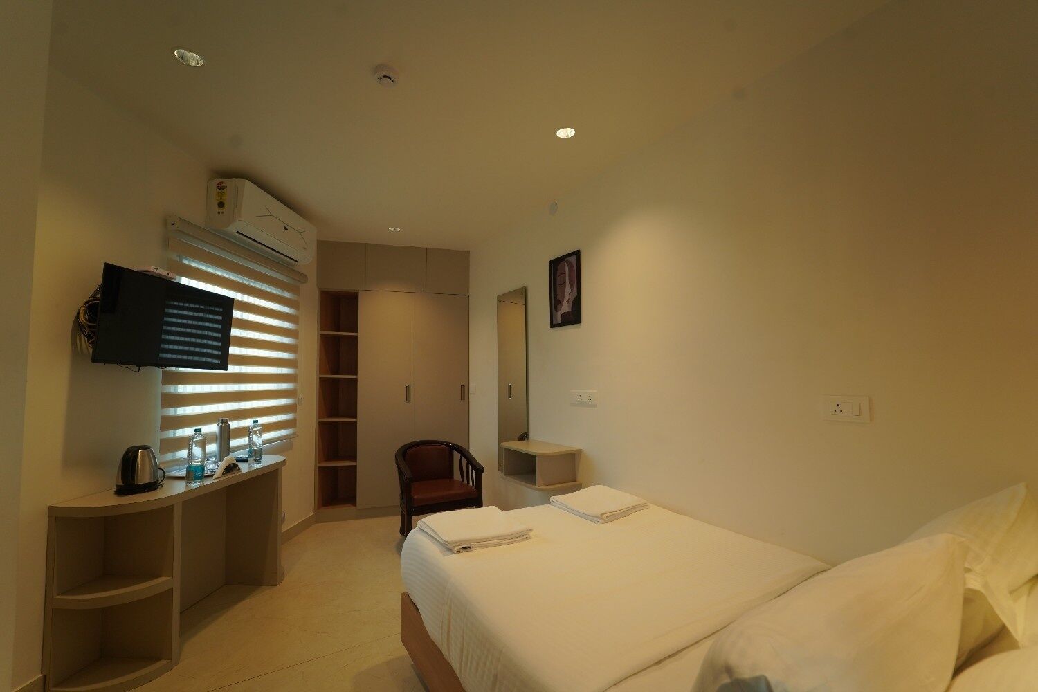 A hotel room with a bed, a desk with a flat-screen TV, a chair, a wardrobe, and a mirror.