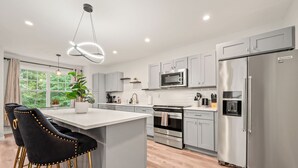 Una cocina moderna con armarios grises, electrodomésticos de acero inoxidable, una isla central con taburetes de bar y un amplio ventanal con vistas a zonas verdes.