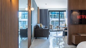 A modern hotel room with a sofa, a TV mounted on the wall, and a balcony with a view of the city. - Mercury Suite Designer Apartment with Pool Access (San Ġiljan)