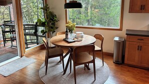 Uma área de refeições com uma mesa redonda, cadeiras, luminária pendente e vista para árvores através de grandes janelas.