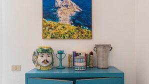 A teal cabinet with a painting of a rocky island, a ceramic jug, a decorative vase, and several books. - Casa Bouganvillea con Terrazza by Wonderful Italy (Isola delle Femmine)