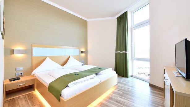 A hotel room with a bed, bedside tables, a TV, and a window with curtains. - Spacious 2-person suite with lake view (Heimbach)