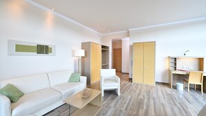 A modern, well-lit room with a white sofa, a small coffee table, a white chair, a desk with a chair, and a wardrobe. - Spacious 2-person suite with lake view (Heimbach)