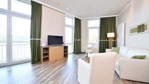 A modern hotel room with a large window, a flat-screen TV on a wooden cabinet, a white sofa, and a lamp. - Spacious 2-person suite with lake view (Heimbach)