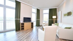 A modern hotel room with a large window, a flat-screen TV on a wooden cabinet, a white sofa, and a lamp. - Spacious 2-person suite with lake view (Heimbach)