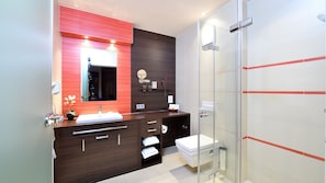 A modern bathroom with a dark wood vanity, a white sink, a glass shower enclosure, and a red accent wall. - Spacious 2-person suite with lake view (Heimbach)