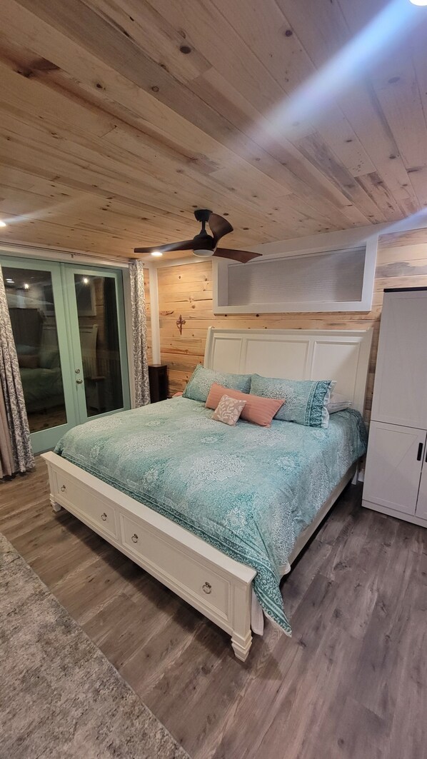A bedroom with a wooden ceiling, a bed with patterned bedding, a ceiling fan, and a sliding glass door.