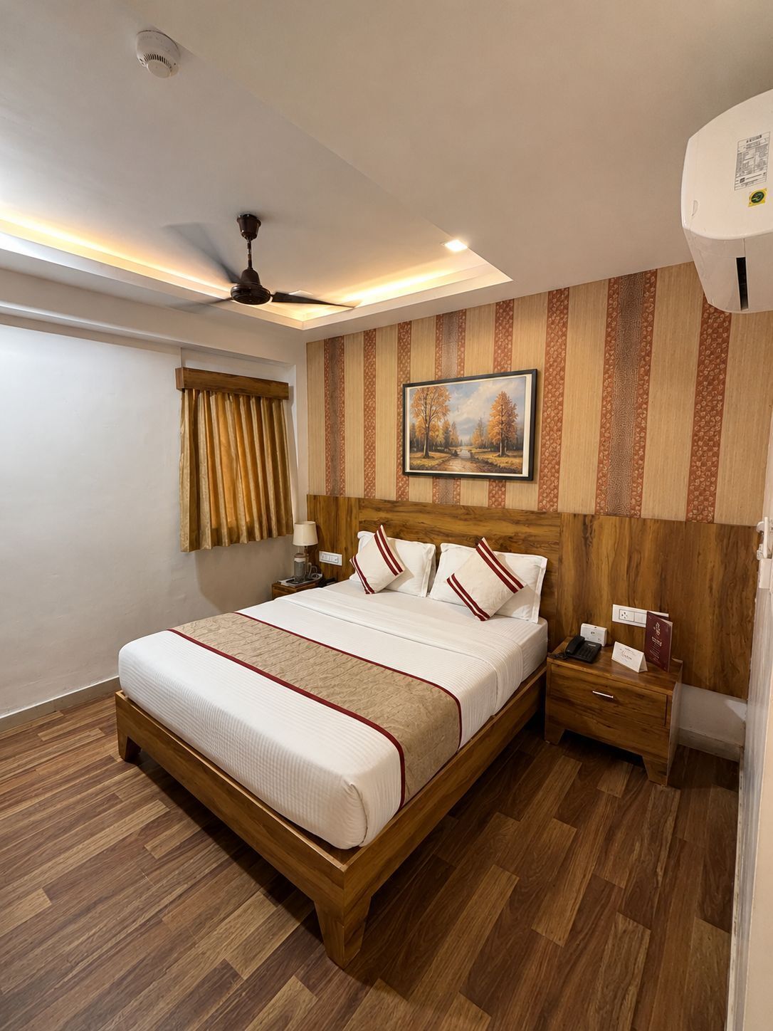 Une chambre d’hôtel bien rangée, avec un lit en bois, une table de chevet, un ventilateur de plafond et un tableau accroché au mur.