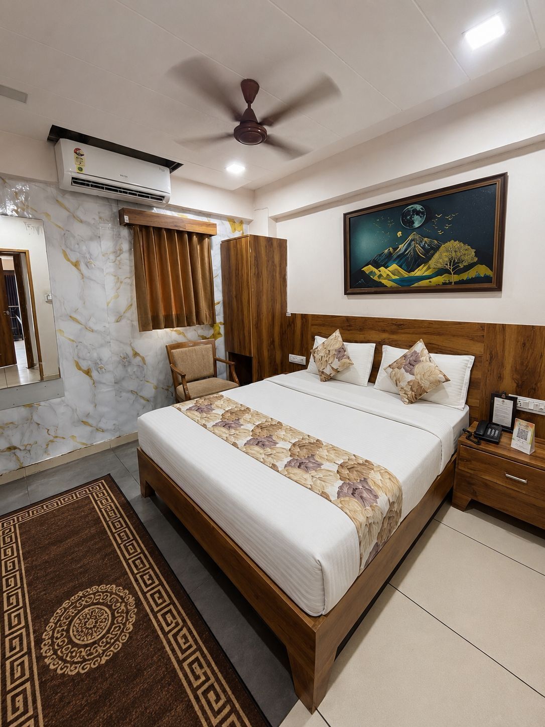 A hotel room with a bed, a nightstand, a desk, a ceiling fan, an air conditioning unit, a painting, and a patterned rug.