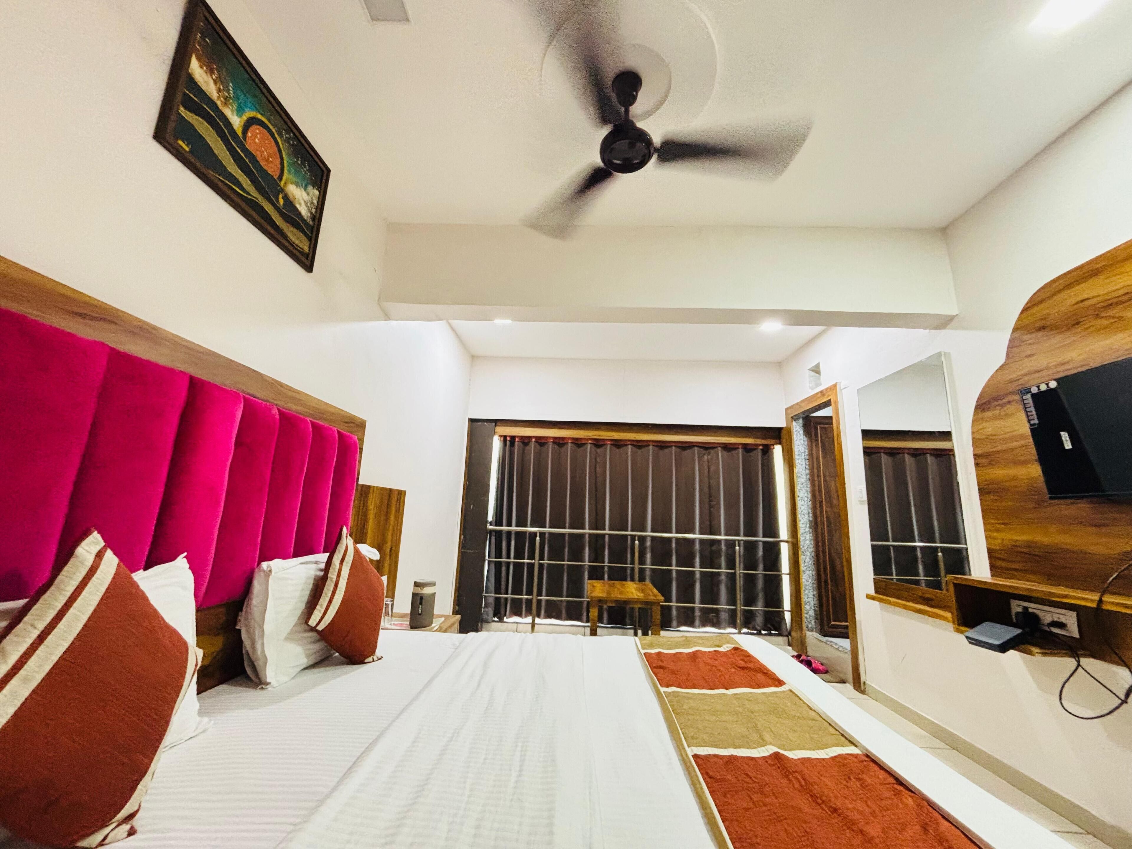 A hotel room with a bed, a ceiling fan, a painting, a window with curtains, a wooden side table, and a TV mounted on the wall.
