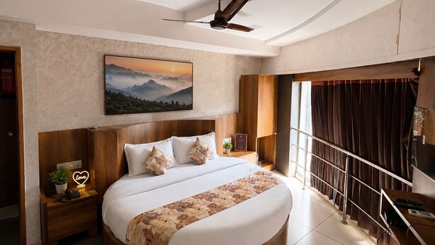 A bedroom with a large bed, a ceiling fan, a painting of mountains, a nightstand with a phone, and a window with curtains. - STAR HOTEL (Ahmedabad)