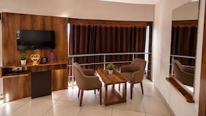 A modern hotel room with a flat-screen TV, wooden paneling, a small desk with a phone, two chairs, a wooden table, and a large mirror. - STAR HOTEL (Ahmedabad)