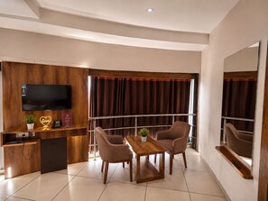 A modern hotel room with a flat-screen TV, wooden paneling, a small desk with a phone, two chairs, a wooden table, and a large mirror.