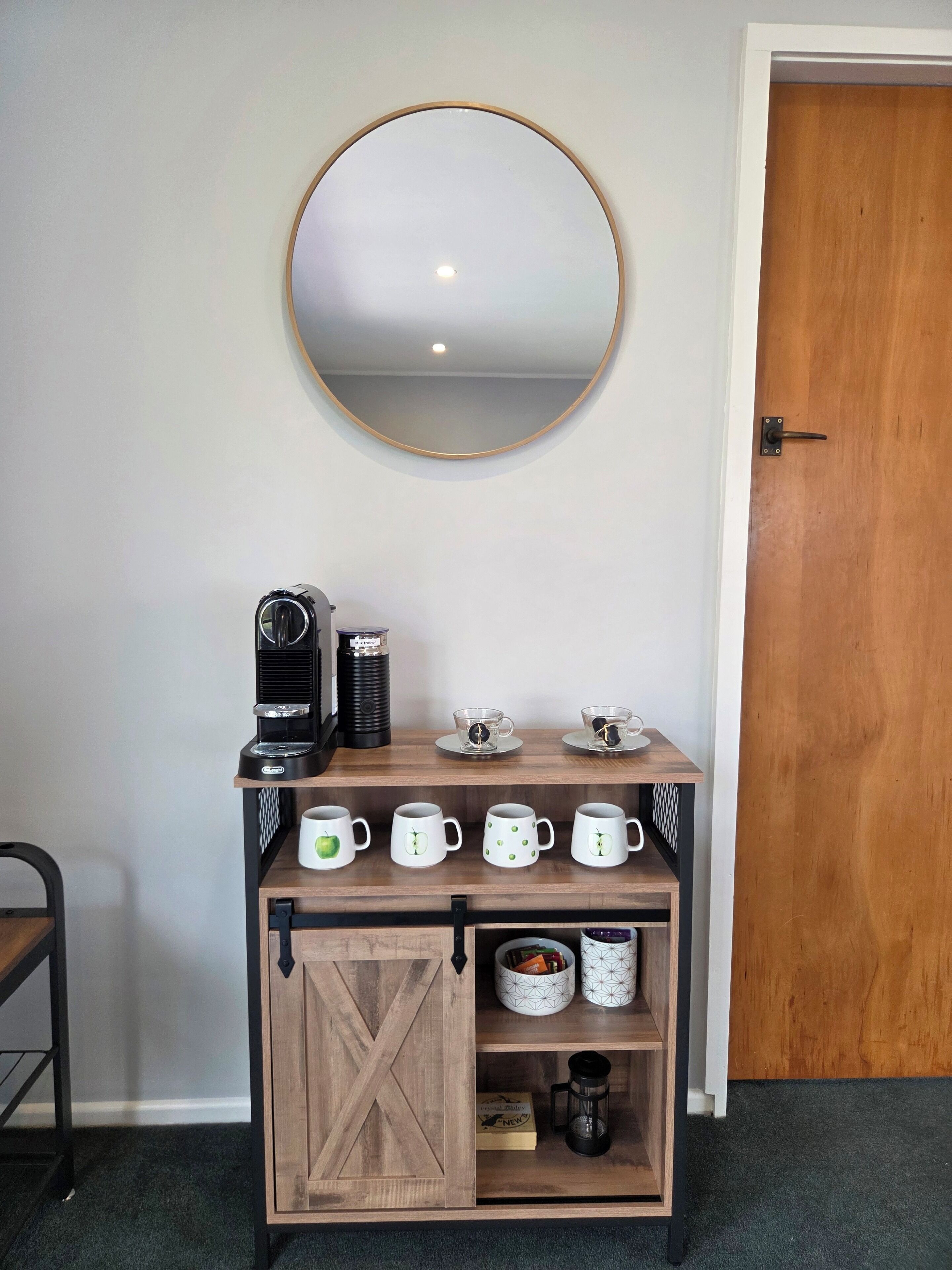 A coffee station with a coffee maker, cups, and mugs on a wooden shelf, a round mirror on the wall, and a wooden door.