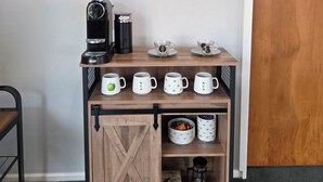 A coffee station with a coffee maker, cups, and mugs on a wooden shelf, a round mirror on the wall, and a wooden door. - Omarama Crib – Spotless Scenic Base for Families (Omarama)