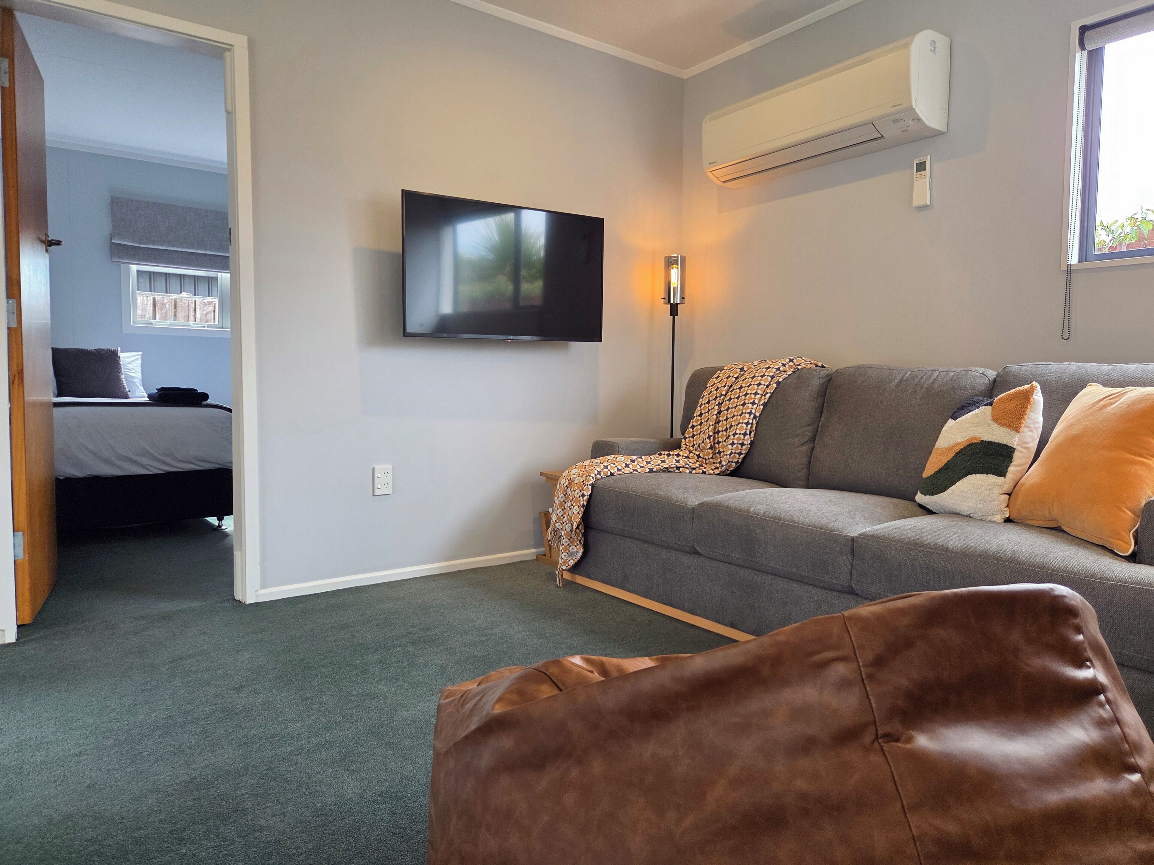 A living room with a grey sofa, a brown leather ottoman, a flat-screen TV mounted on the wall, and a window with blinds.