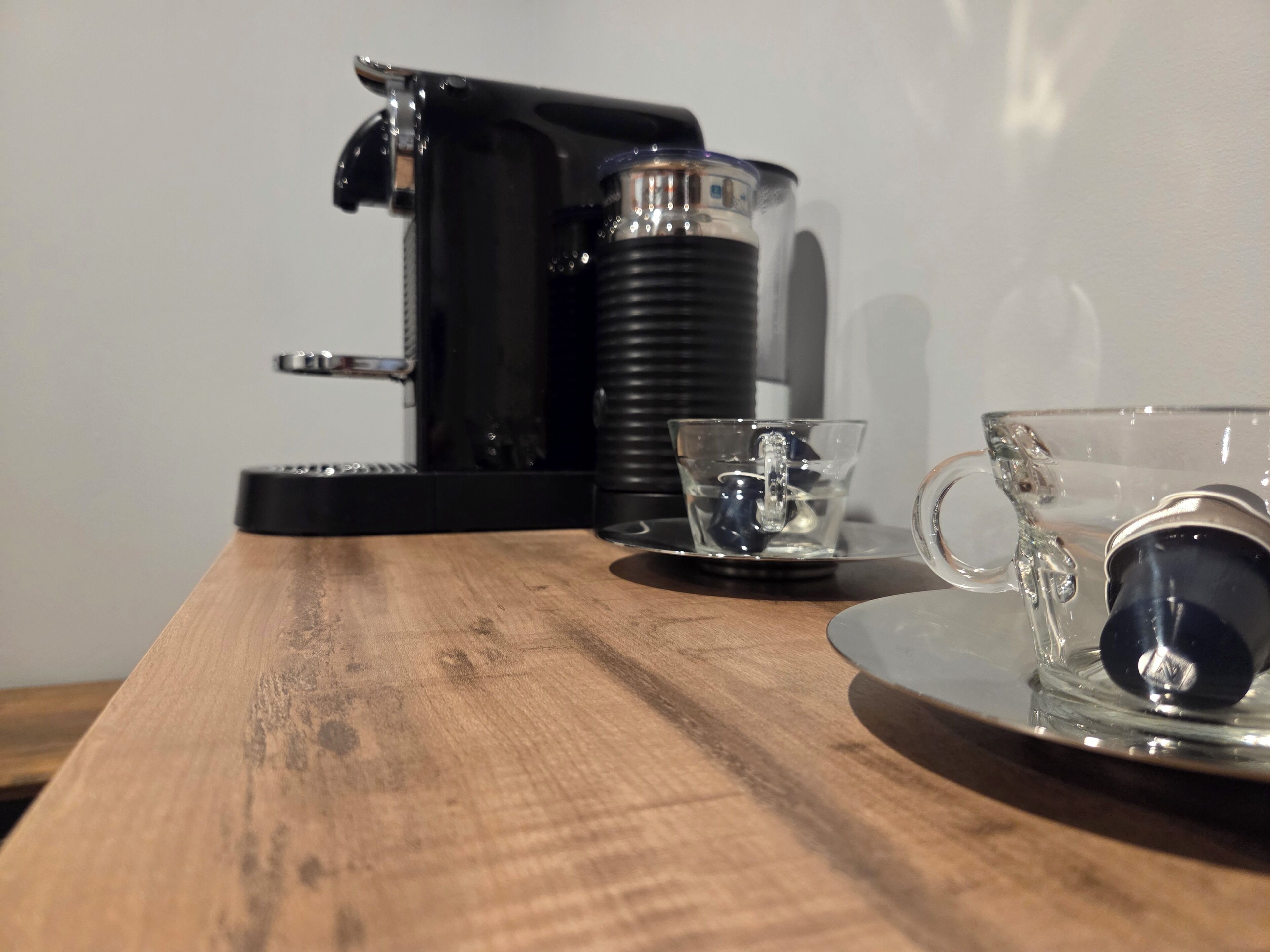 A coffee maker, a glass cup, and a saucer on a wooden table.