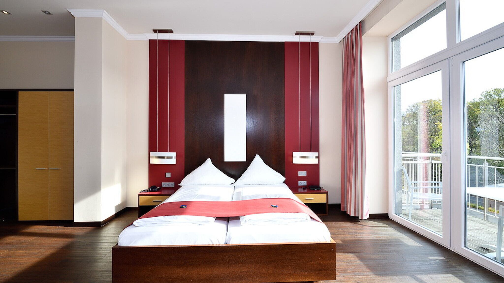 Quarto de hotel moderno com uma cama grande, cabeceira de madeira, duas mesas de cabeceira com luminárias, uma janela ampla com vista para o exterior e uma parede de destaque vermelha.
