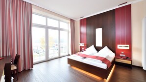A modern hotel room with a large bed, a desk, and a view of the city through large windows. - Double room Seeblick with balcony (Heimbach)