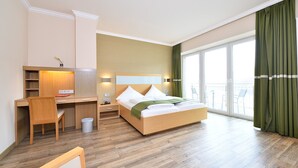 A hotel room with a wooden bed, a desk, a chair, a small trash bin, and a balcony with green curtains. - Double room Seeblick with balcony (Heimbach)