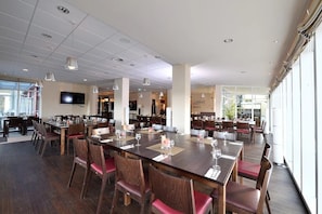 A spacious dining area with multiple tables and chairs, a flat-screen TV mounted on the wall, and large windows providing natural light.