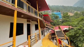 A multi-story building with a red roof and yellow railings. There are multiple windows and a staircase leading to the upper floors. The building is surrounded by greenery and hills in the background. - New Mandakini River Resort (Ukhimath)