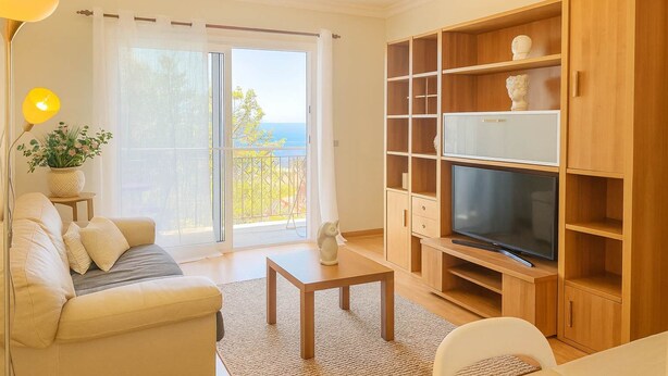 A living room with a sofa, a coffee table, a TV, wooden shelves, and a balcony with a view. - Titou Apartment Sea View (Machico)