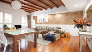 Uma sala de estar moderna com uma mesa de jantar de vidro, piso de madeira, parede de tijolos decorativa e um sofá com almofadas estampadas.