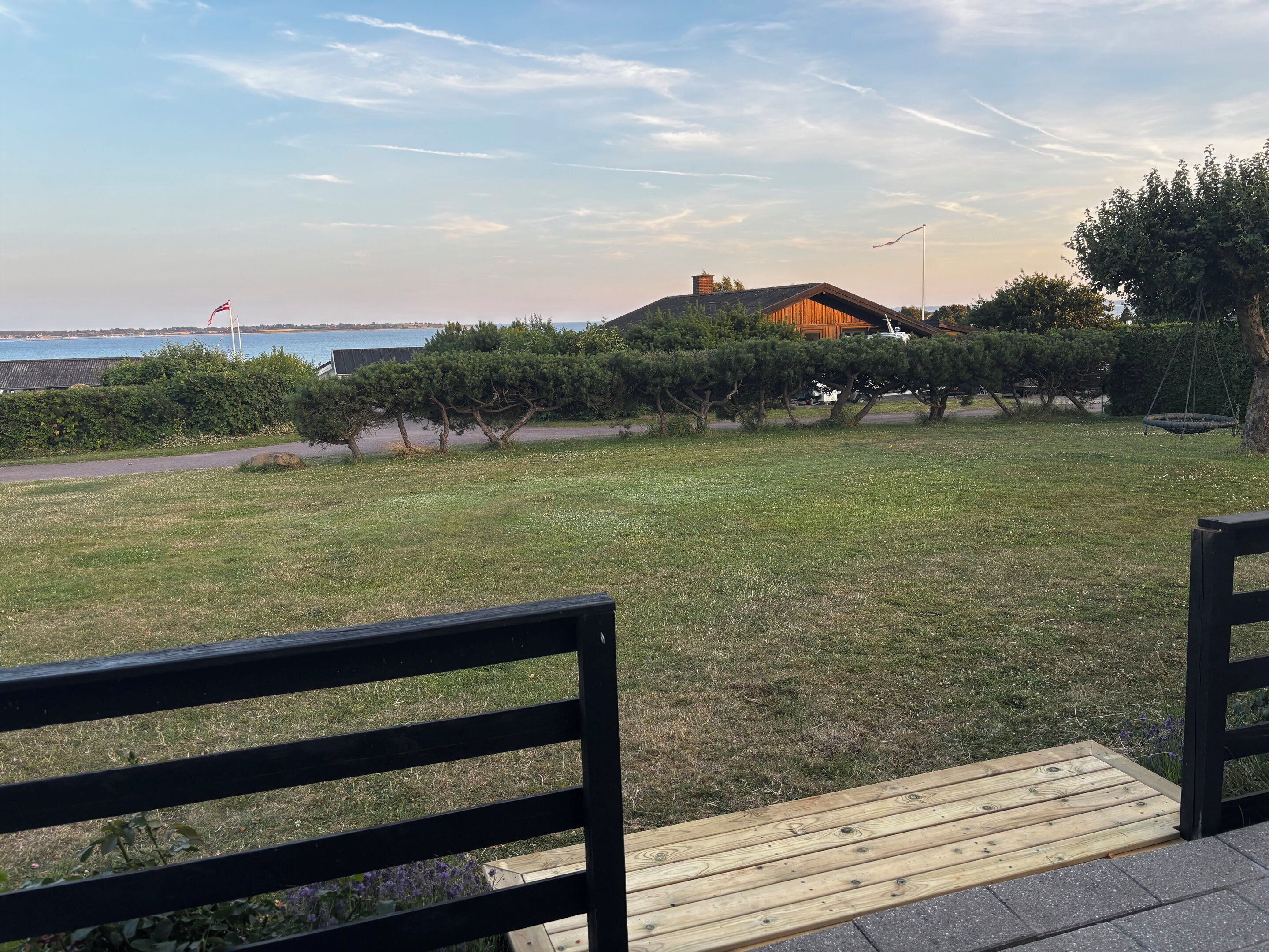 Un espacio al aire libre con césped, una terraza de madera y un banco, una bandera y un edificio con techo inclinado al fondo.