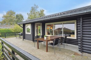 A log cabin with a patio area featuring a wooden table and chairs, large windows, and a view of the surrounding landscape.