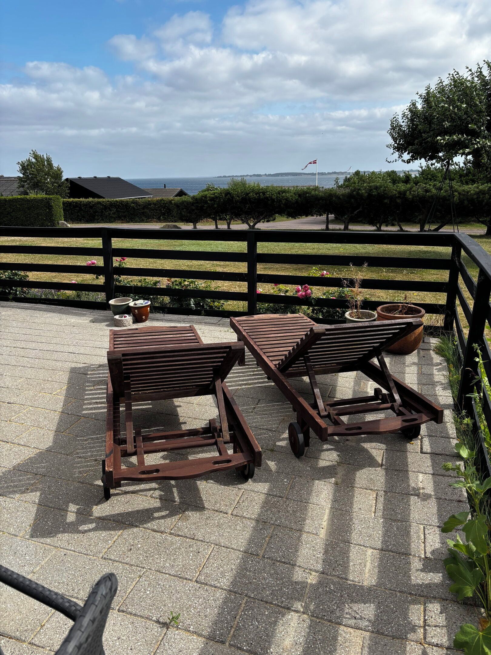 A patio with two wooden lounge chairs and a table, overlooking a landscaped area with a view of the sea and distant hills.