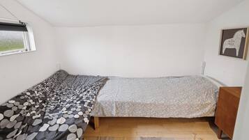 A small, single-bed room with a window, a wooden dresser, and a framed picture on the wall.