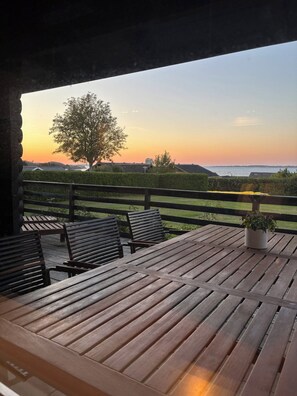 An outdoor seating area with wooden tables and chairs, a potted plant, and a view of a tree and landscape during sunset.