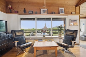 A modern living room with a wooden ceiling, large windows, two leather chairs, a wooden table, and a television.