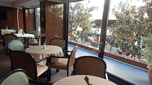 Um restaurante com mesas e cadeiras redondas, grandes janelas com vista para a rua, e um vaso com uma flor em uma das mesas.