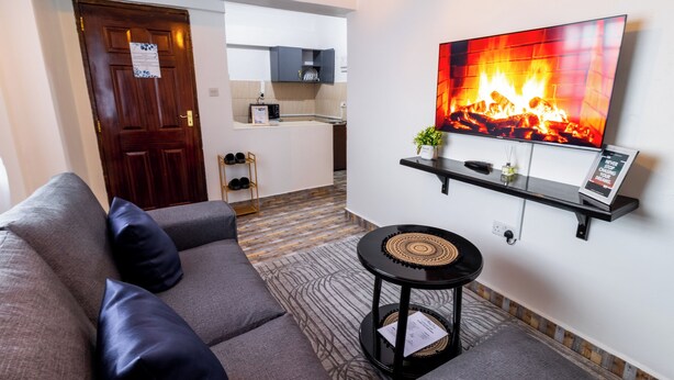 Ein Wohnzimmer mit grauem Sofa, einem kleinen schwarzen Beistelltisch und einem an der Wand befestigten Fernseher, der einen Kamin zeigt. Im Hintergrund ist eine Küche mit Mikrowelle und Schränken zu sehen.