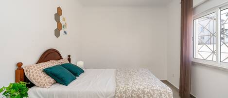 Un dormitorio con una cama de madera, una mesita de noche con una planta, una ventana con cortinas y un adorno en la pared.