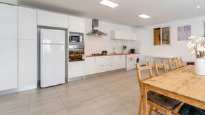 Een moderne keuken met witte kasten, een roestvrijstalen koelkast, een magnetron en een gasfornuis. Er staat een eettafel met stoelen en een bloevas.
