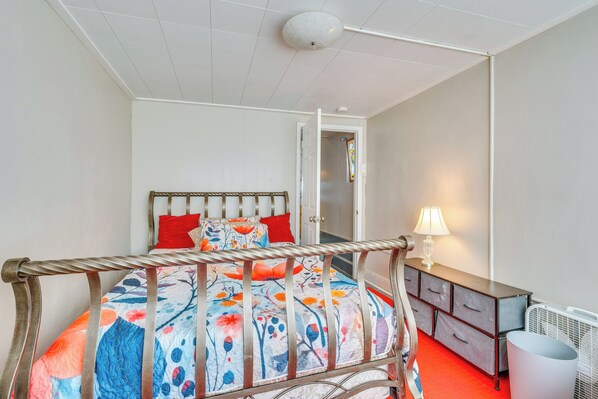 Une chambre à coucher avec un lit en métal, une couvre-lit à motifs floraux colorés, un oreiller rouge, une table de chevet avec une lampe et un radiateur.