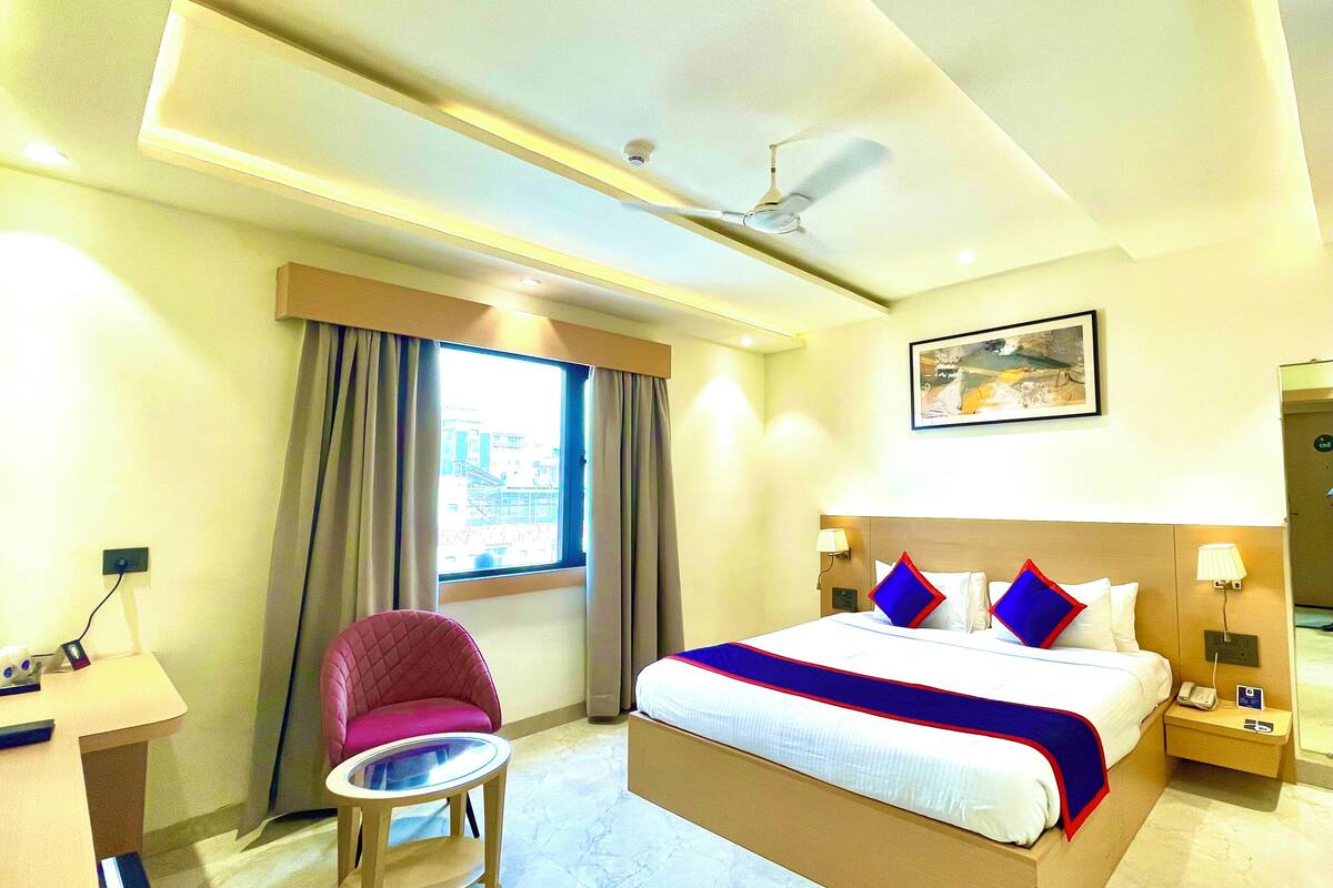 A hotel room with a bed, a desk, a chair, a nightstand, a lamp, a ceiling fan, and a window with curtains.