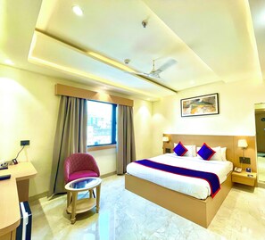 A hotel room with a bed, a desk, a chair, a nightstand, a lamp, a ceiling fan, and a window with curtains.