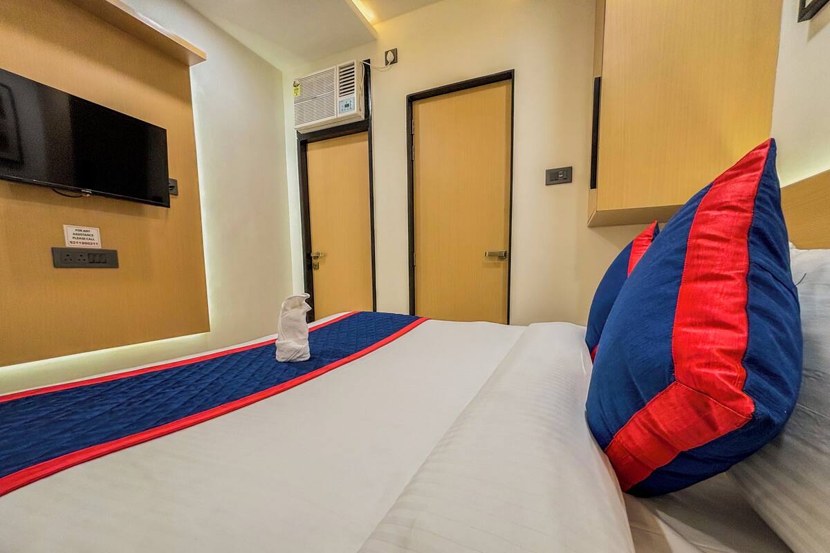 A hotel room with a bed, a flat-screen TV mounted on the wall, an air conditioning unit, and a water bottle on the bed.