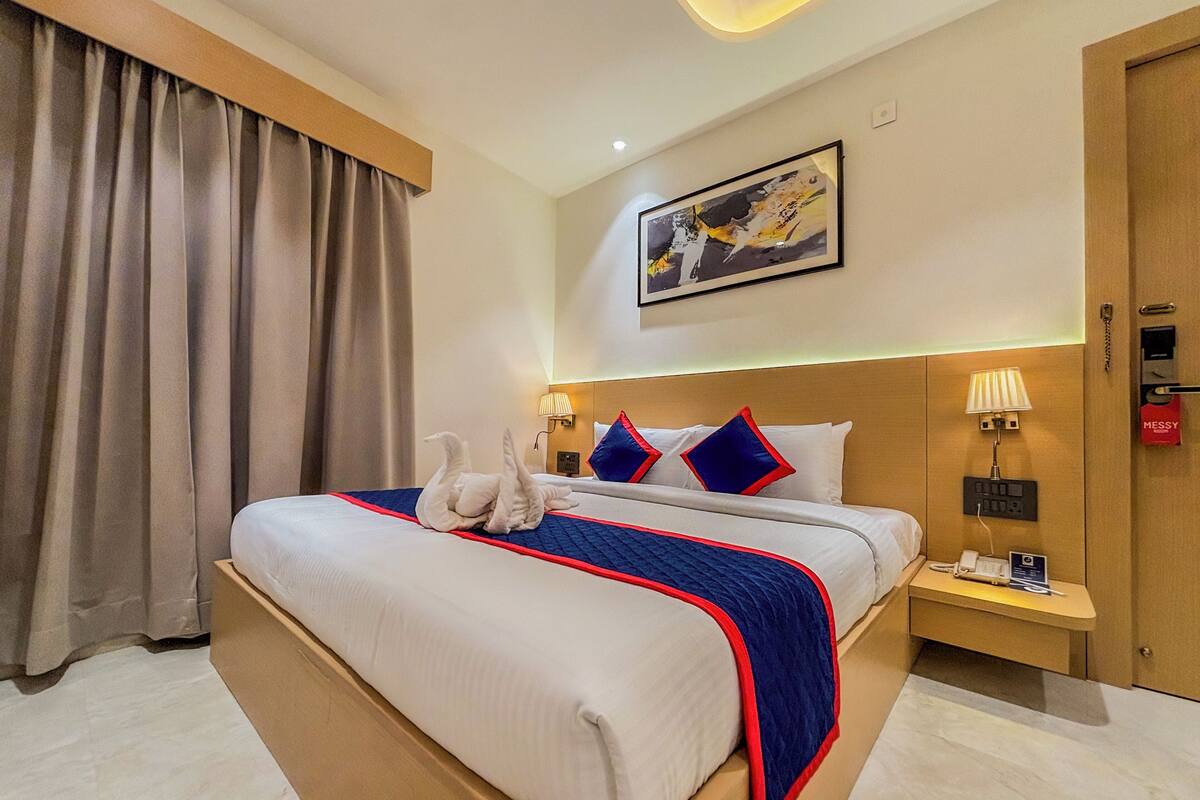 A hotel room with a large bed, a nightstand, a lamp, a painting on the wall, and a door with a 'MESSY' sign.