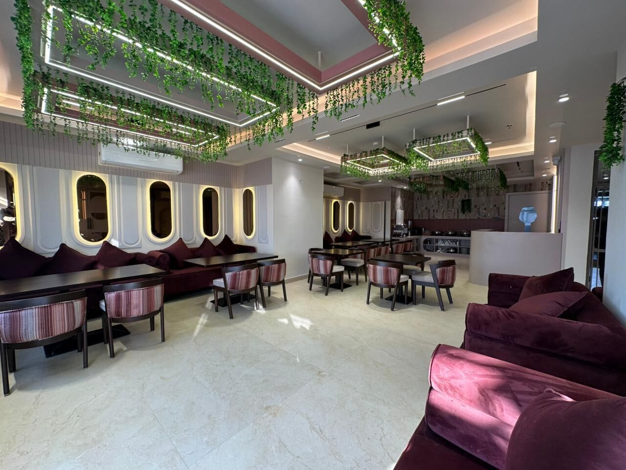 A modern interior with a seating area featuring a sofa and chairs, a dining area with tables and chairs, and a ceiling adorned with green plants and lighting.