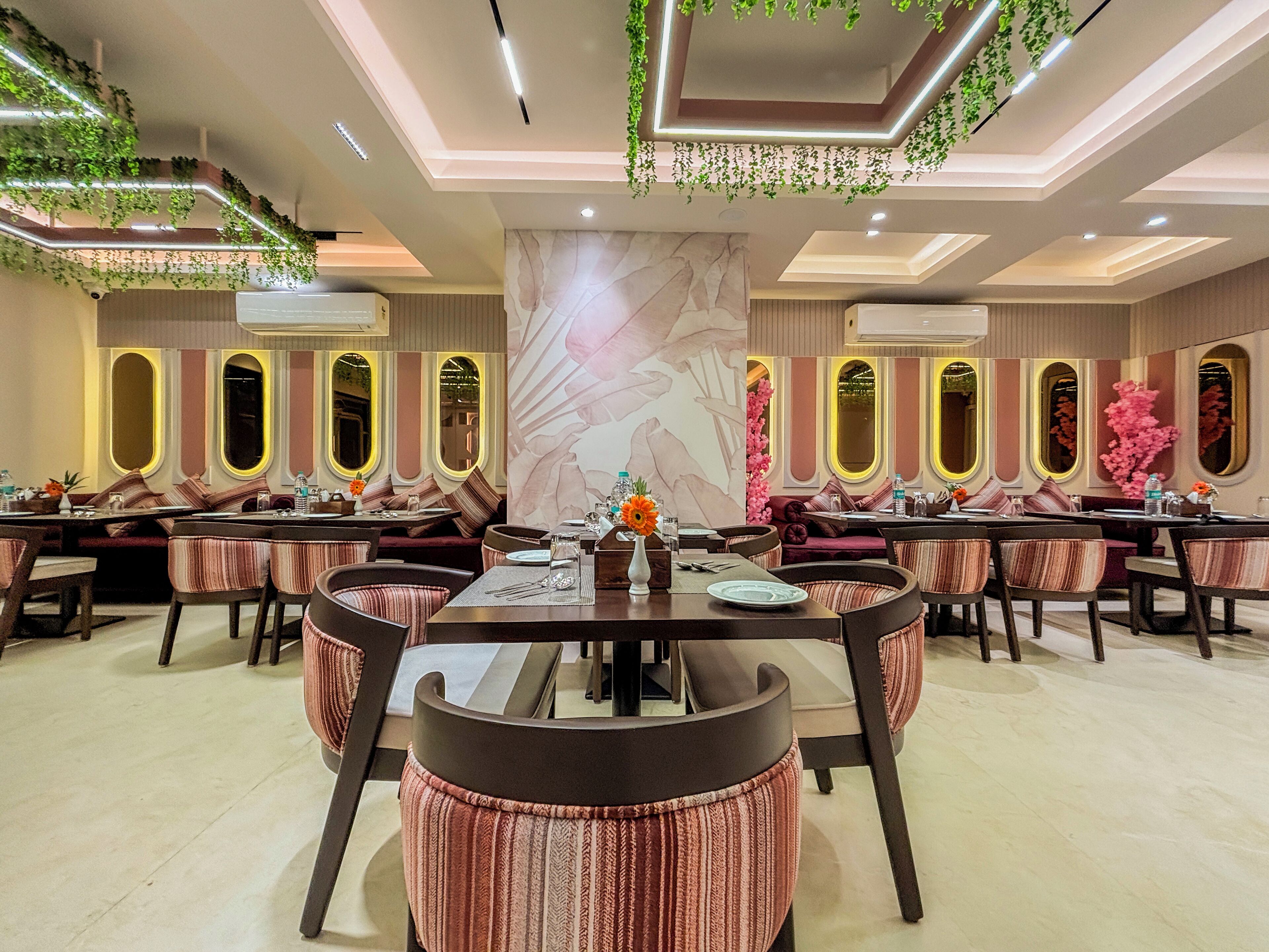 A modern dining area with multiple tables and chairs, decorative plants, and a large floral mural on the wall.
