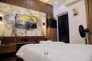 Ein modernes Hotelzimmer mit einem großen Fernseher an einer Marmorwand, einem Bett mit weißer Bettwäsche, einem Holzkonsolentisch und einem schwarzen Ventilator.