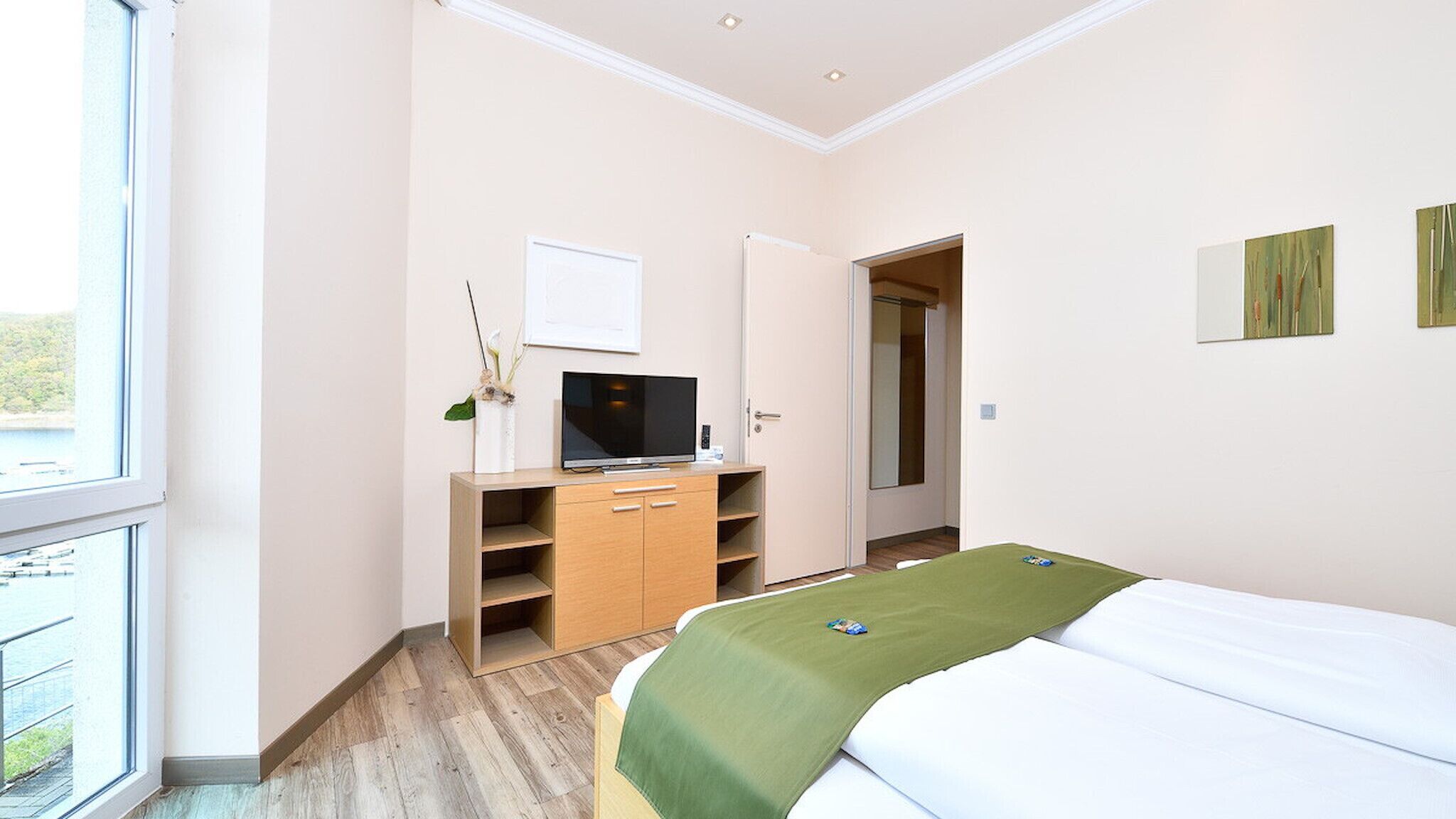 Habitación de hotel con cama, un televisor sobre un mueble de madera, una puerta que da a otra habitación y una ventana con vistas.