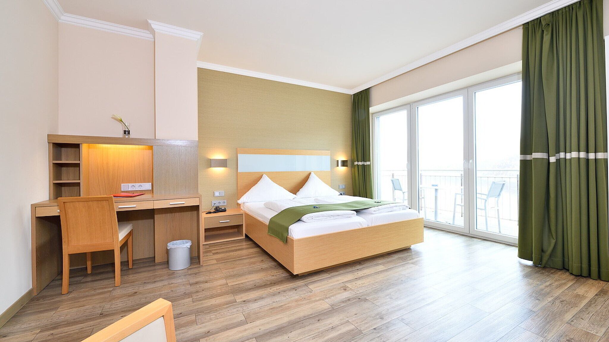 Hotelkamer met een houten bed, een bureau, een stoel, een kleine prullenbak en een balkon met groene gordijnen.